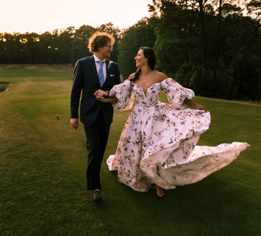 Bride in floral gown and groom walking together on a golf course at sunset